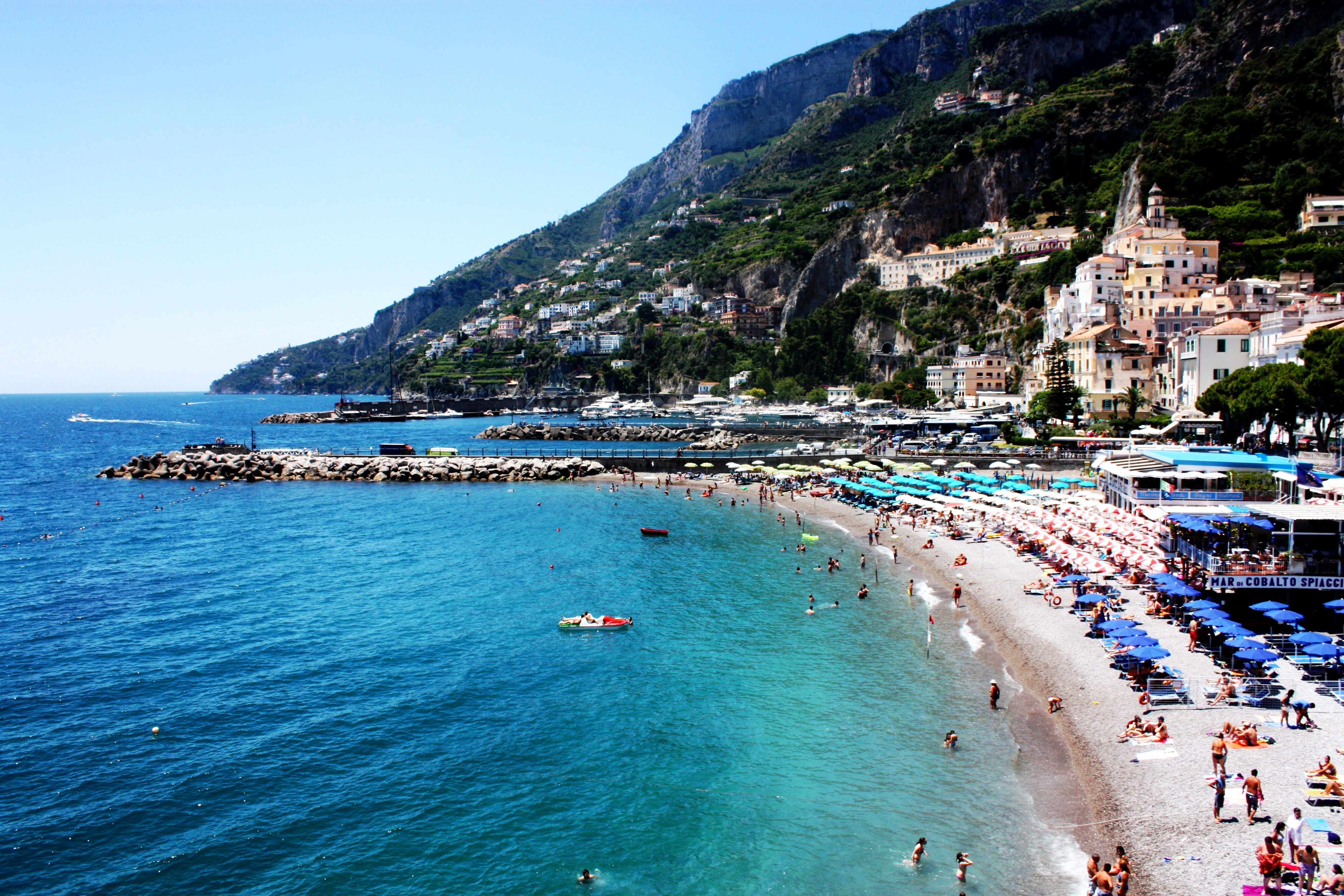 Amalfi beach and waterfront below steep green hills, with boats in the harbour