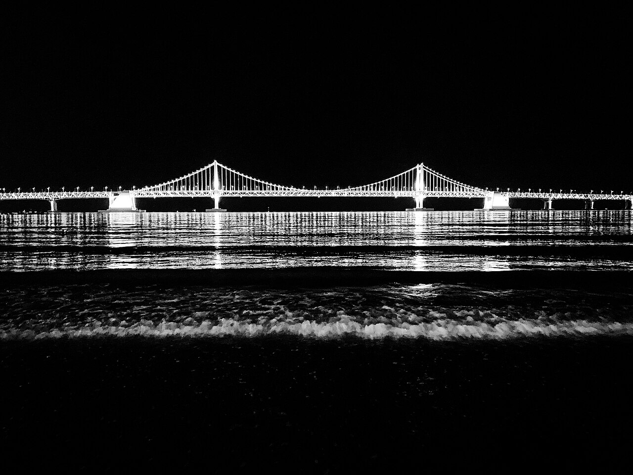 Black-and-white night photograph of the illuminated Gwangandaegyo bridge above waves at Busan's Gwangalli Beach