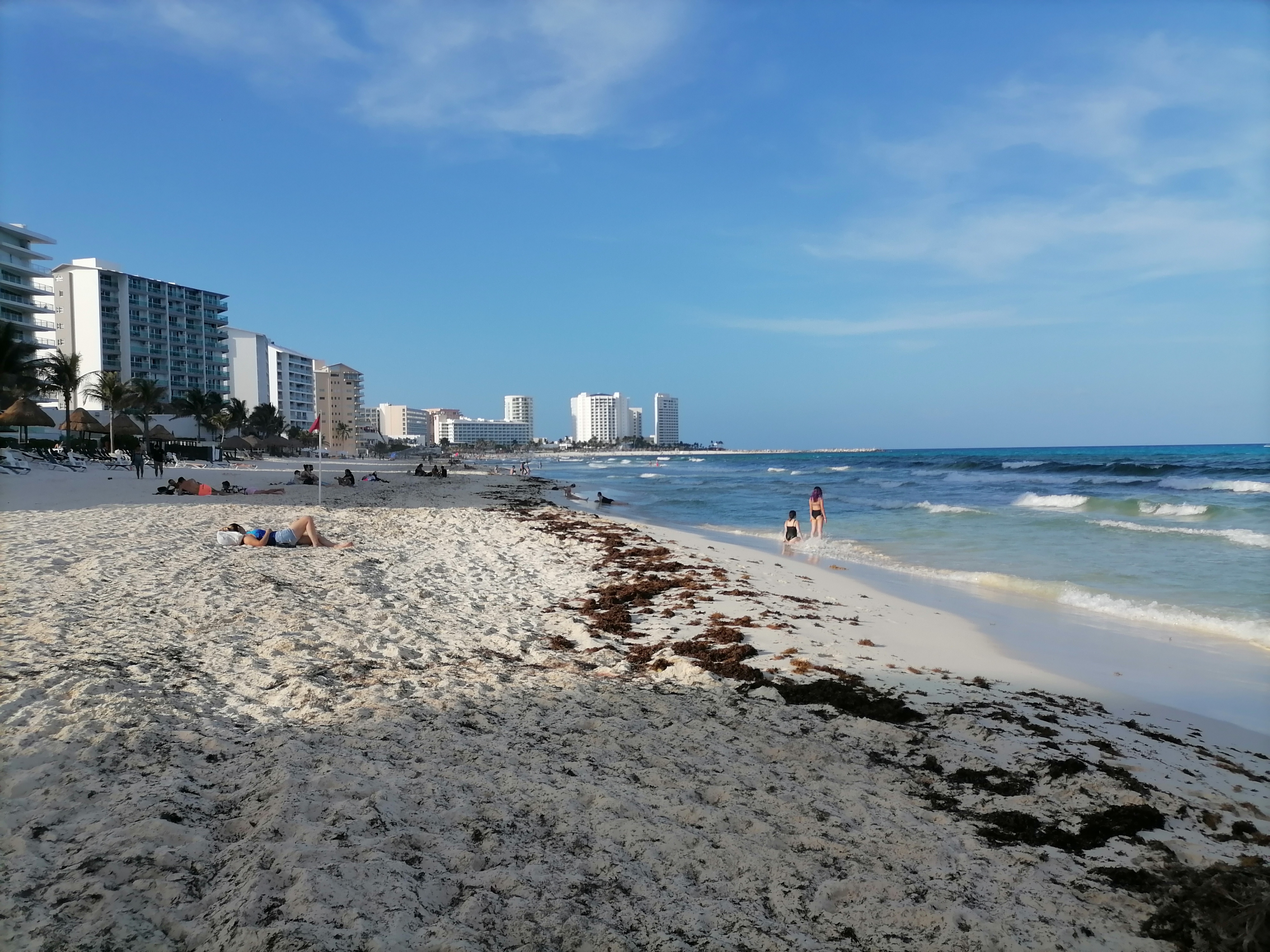 Cancún shoreline with pale sand, seaweed along the water, beachgoers and hotel buildings beside the blue sea