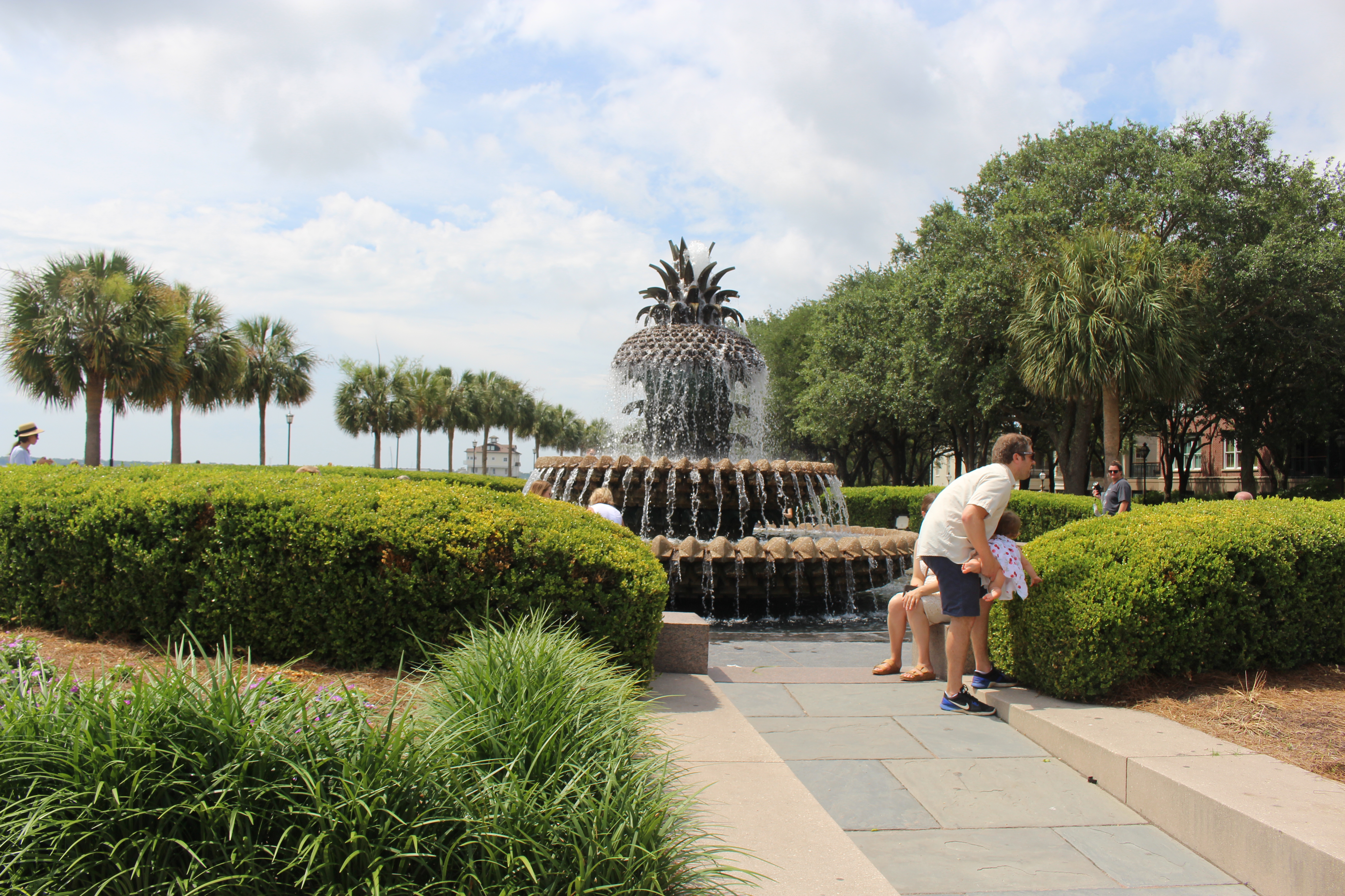 Pineapple-shaped fountain spilling water into a stone basin, with hedges and palms around it and visitors on the paved path