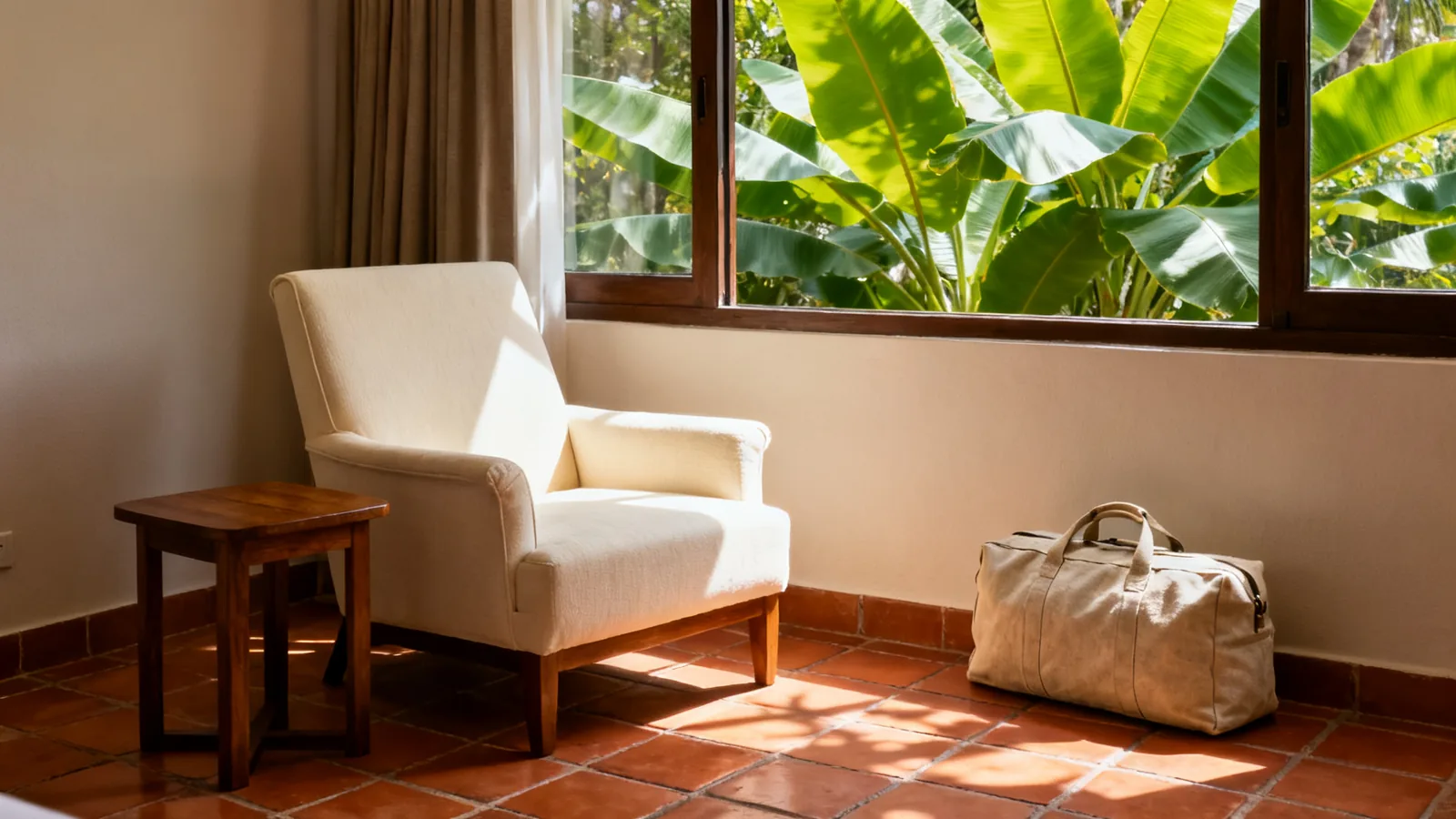 A cream hotel reading chair beside a timber table and canvas travel bag, with green leaves outside