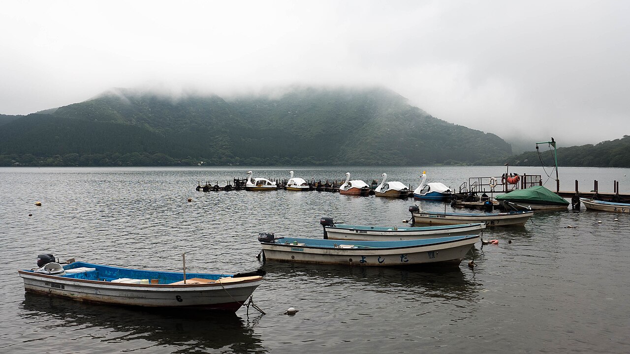 Small boats and swan-shaped pedal boats moored on Lake Ashi beneath mist-covered hills