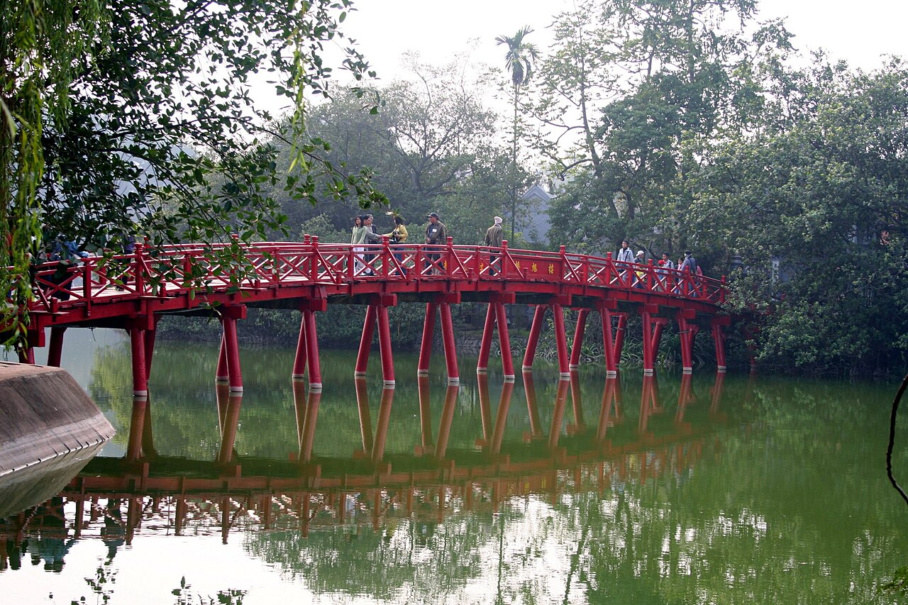 The red The Huc Bridge reflected in Hoan Kiem Lake in Hanoi, framed by green trees