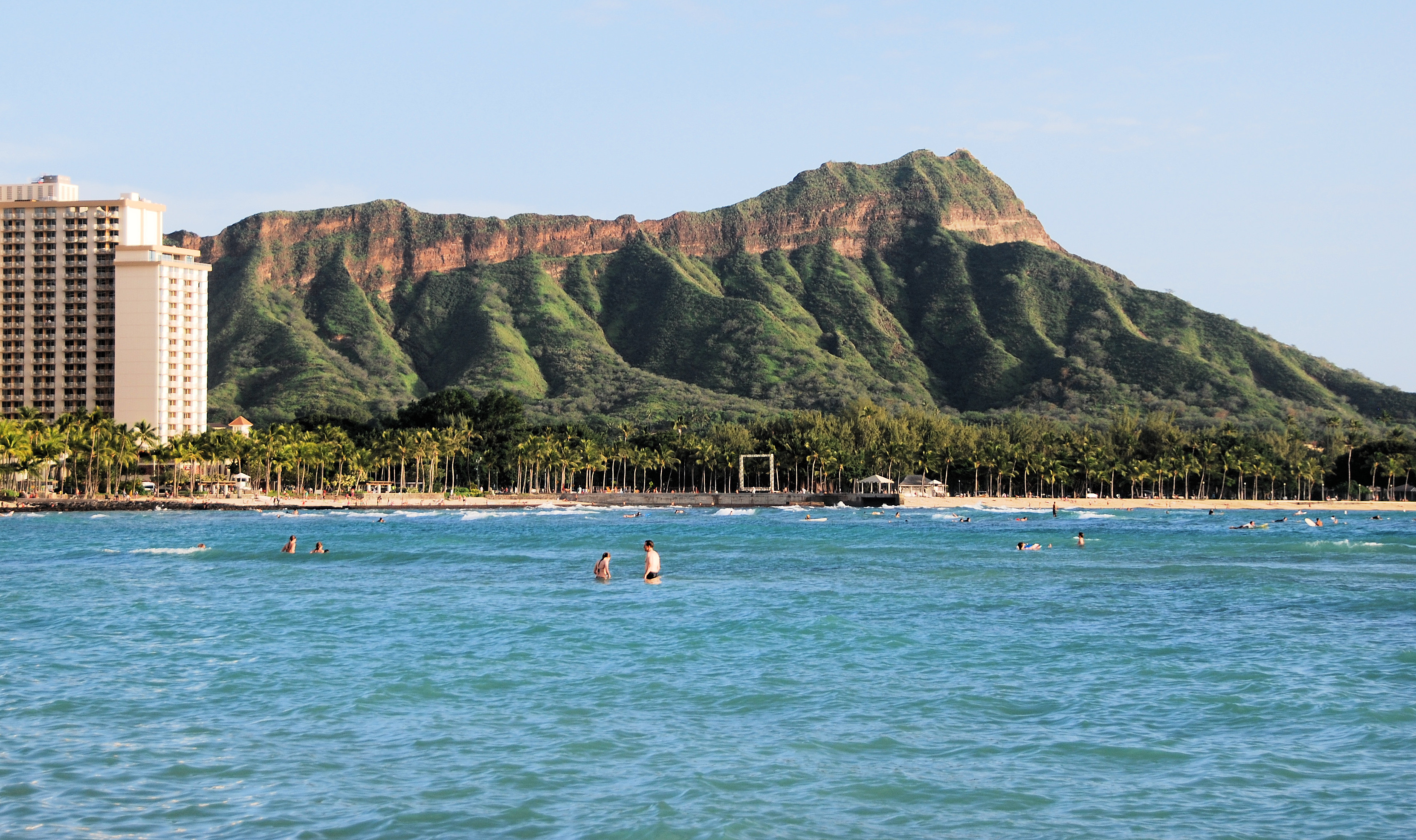 Blue ocean with swimmers before a palm-lined shore, a tall building at left and green Diamond Head ridges behind