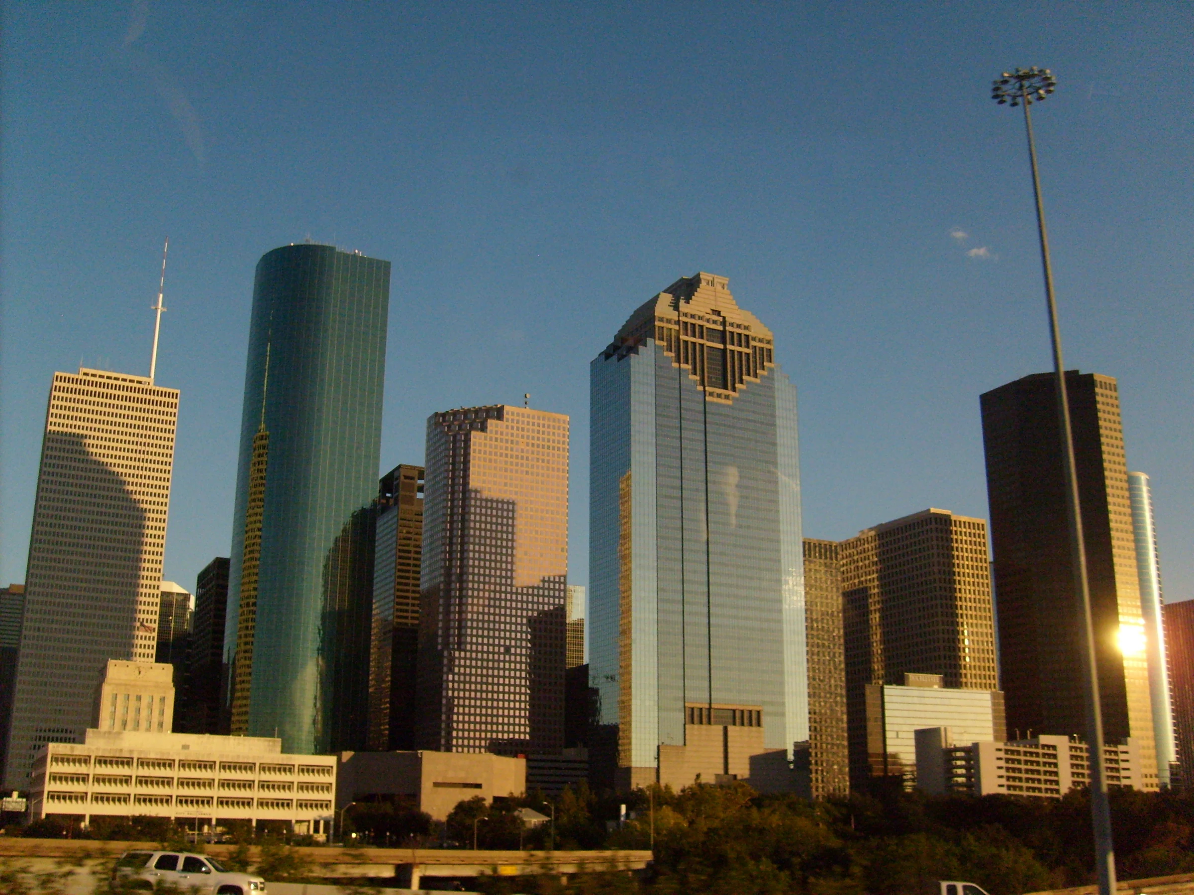 Warm-lit Downtown Houston towers beneath blue sky with a road and tall light pole in the foreground