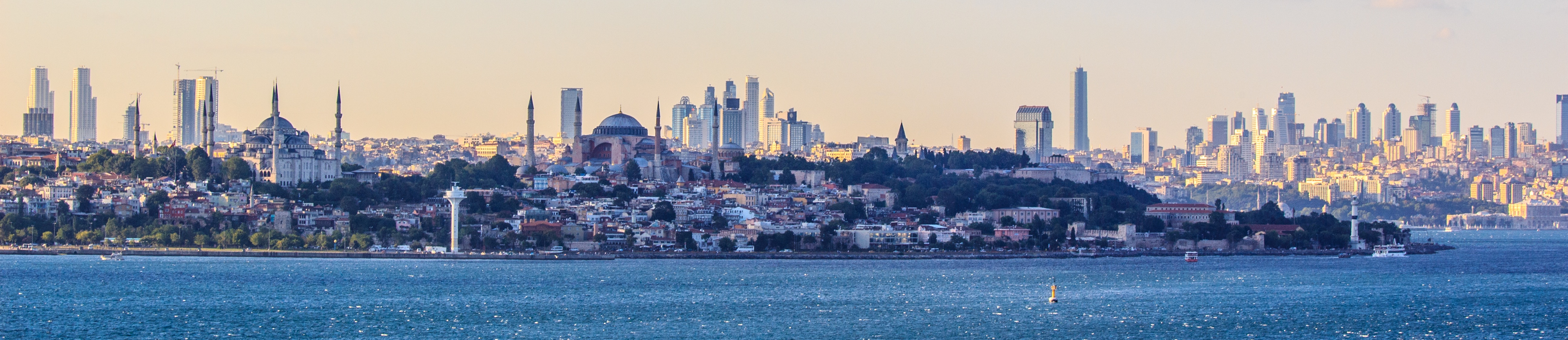 Historic domes and dense rooftops backed by high towers, with blue water in front beneath a hazy sky