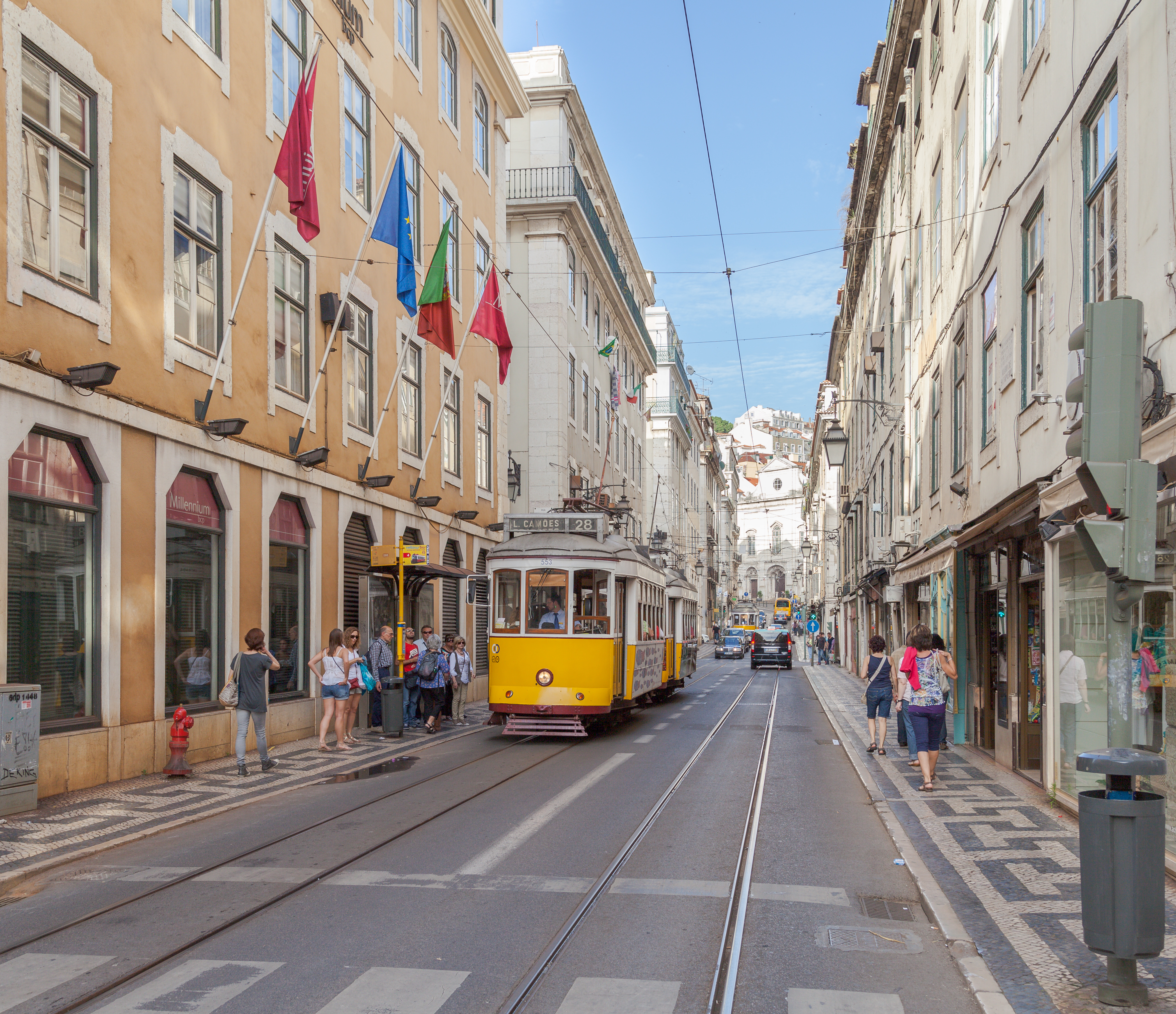 A yellow Lisbon tram on Rua da Conceição between tall buildings, with people on patterned pavements and rails leading along the street