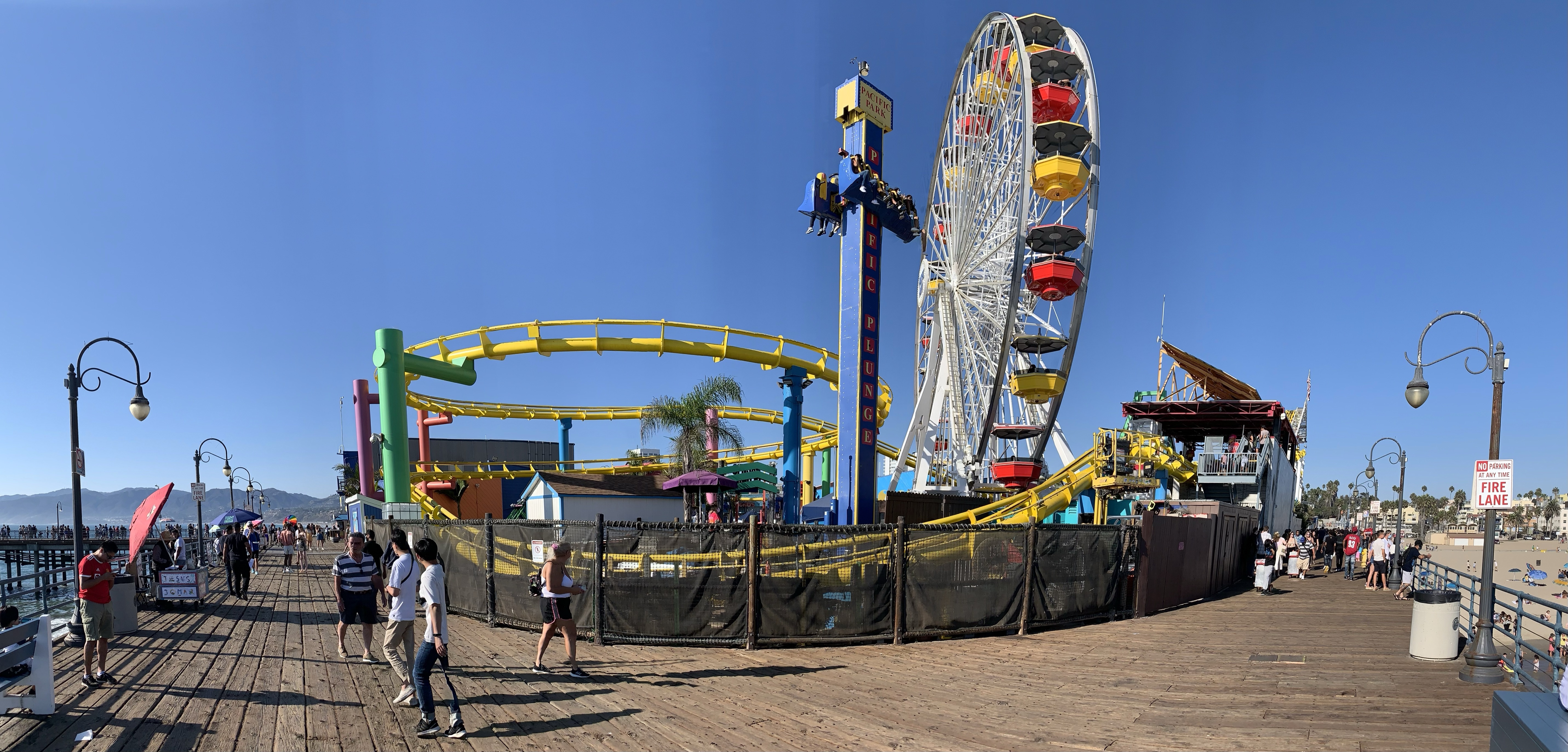 Ferris wheel and yellow roller-coaster track beside the wooden Santa Monica Pier boardwalk, with visitors beneath a blue sky