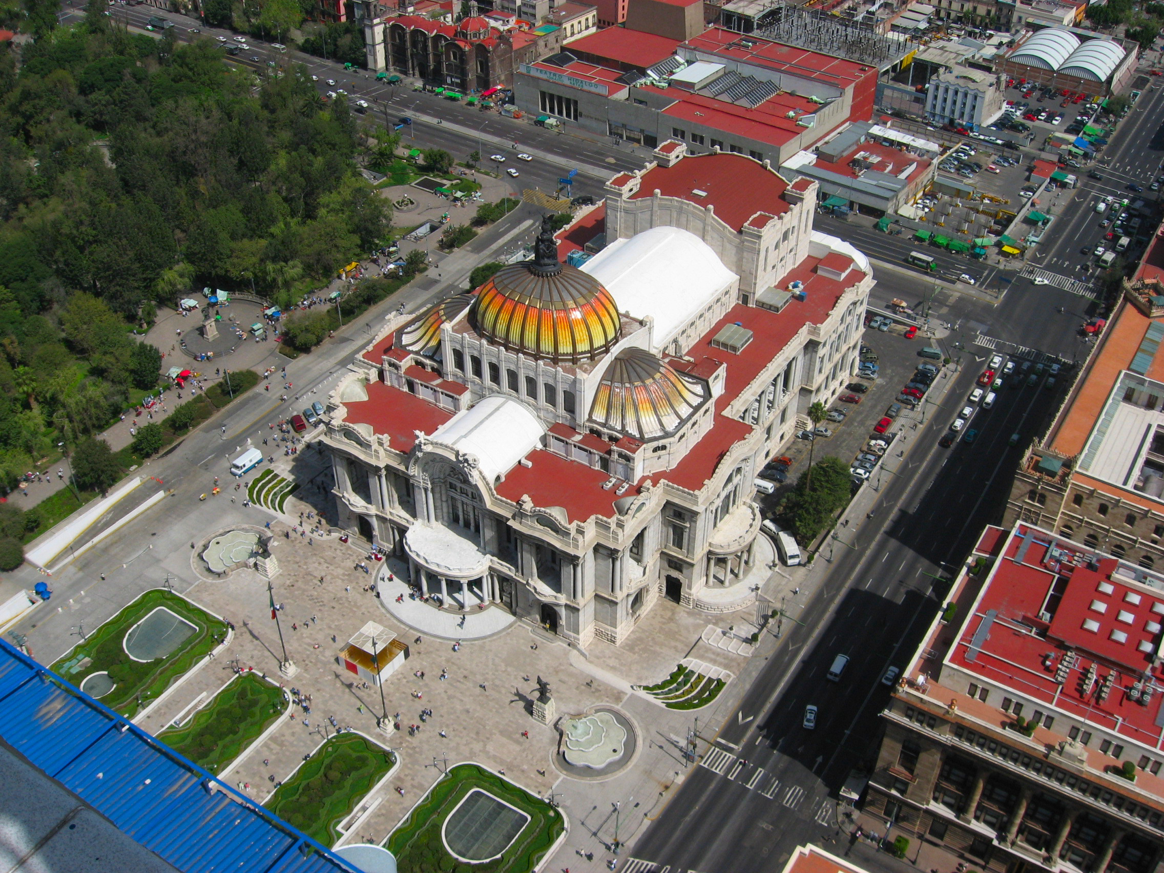 Palacio de Bellas Artes seen from above, with coloured domes, pale stone façades, nearby streets and people in the forecourt