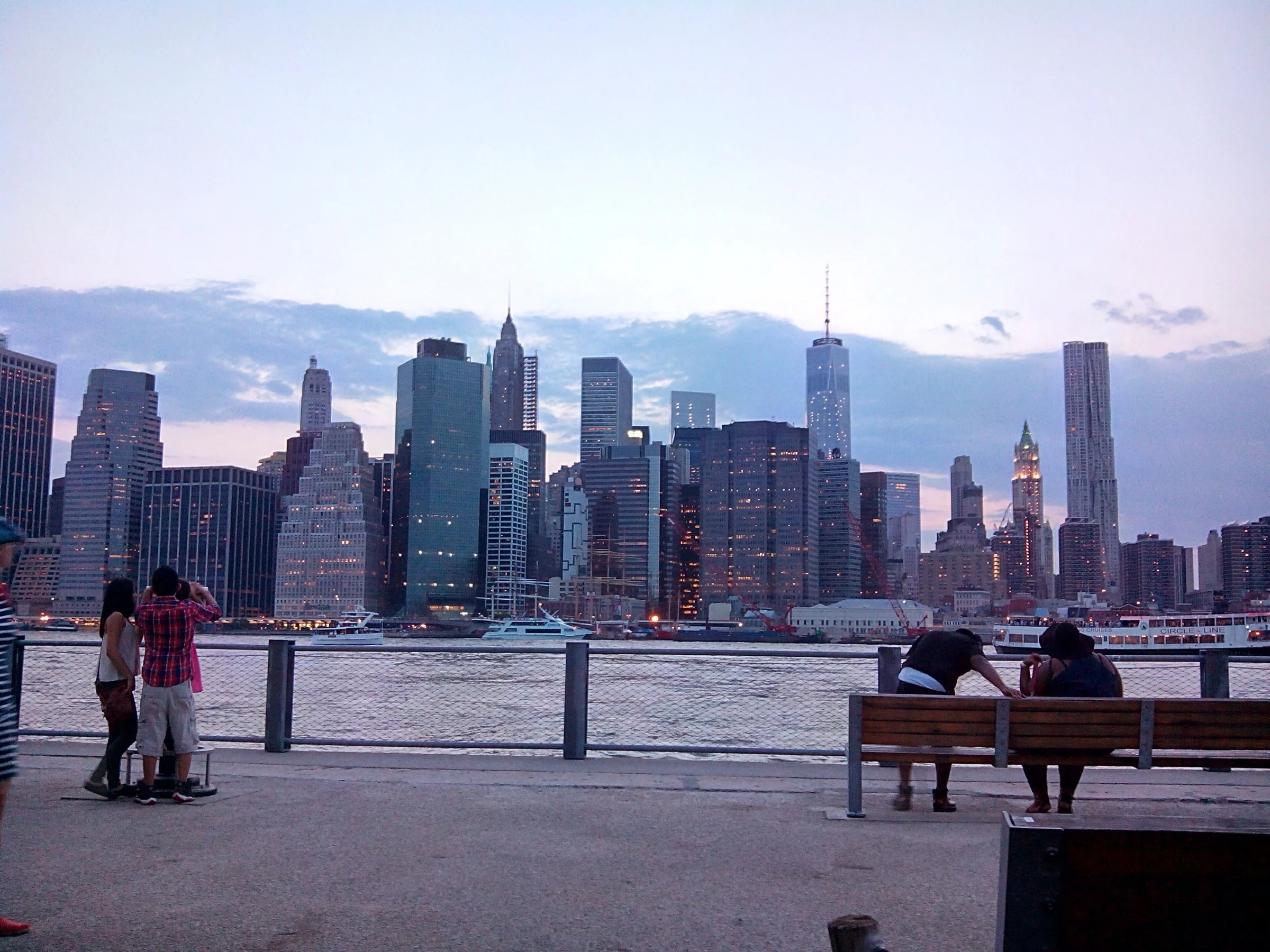 Historical Lower Manhattan skyline across the East River from Brooklyn Bridge Park with a waterfront railing and visitors