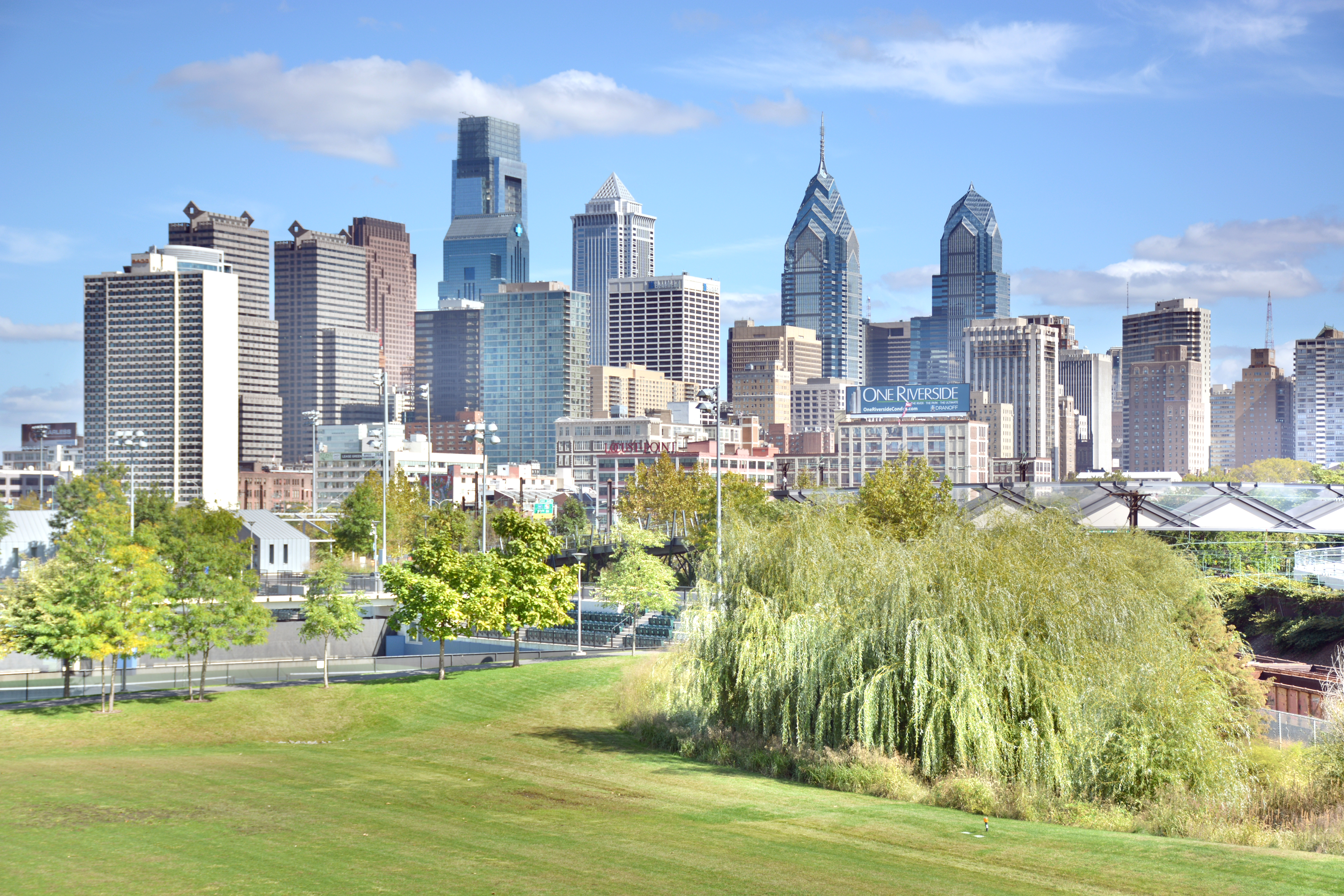 Philadelphia towers under blue sky, with green lawns and leafy trees in the foreground