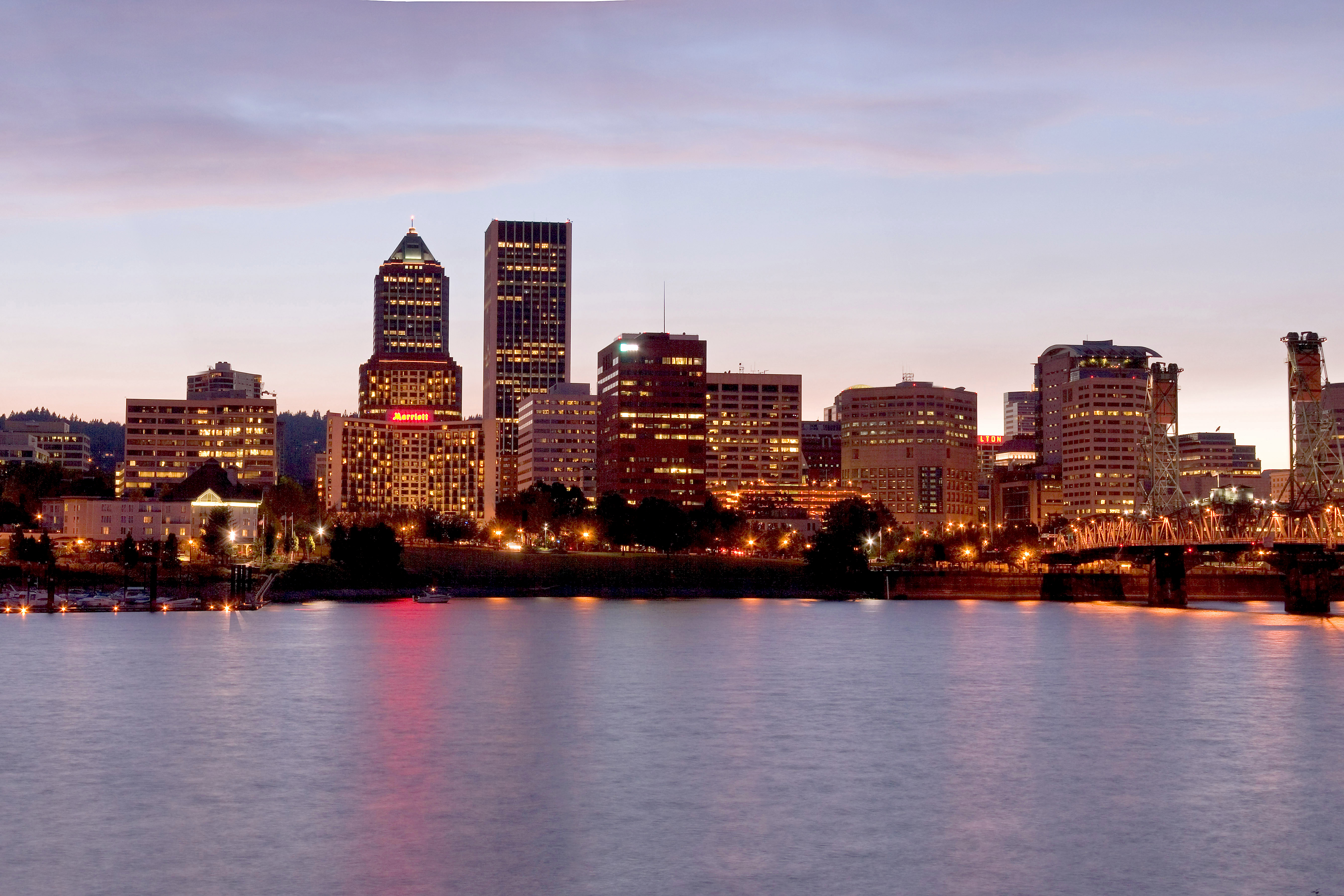 Lit Portland skyline above a broad river at dusk, with reflections on the water and a metal bridge at right