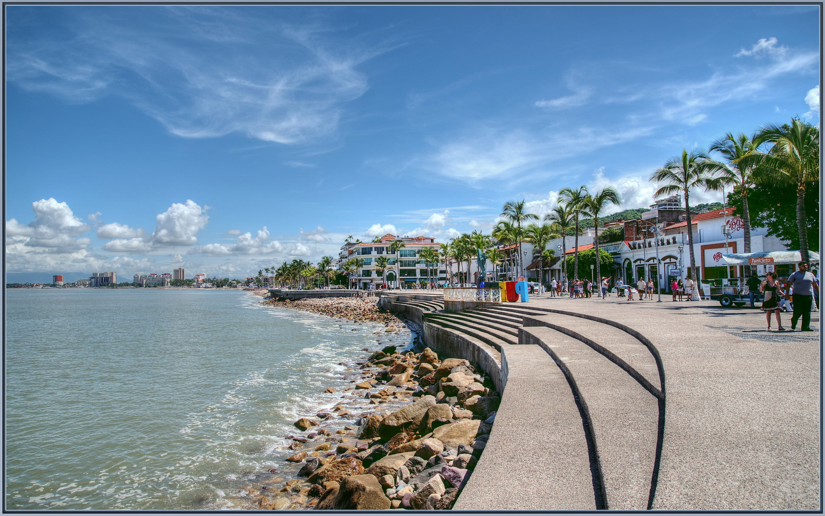 Curved steps beside the water on Puerto Vallarta’s Malecón, with palms, seafront buildings and people on the promenade