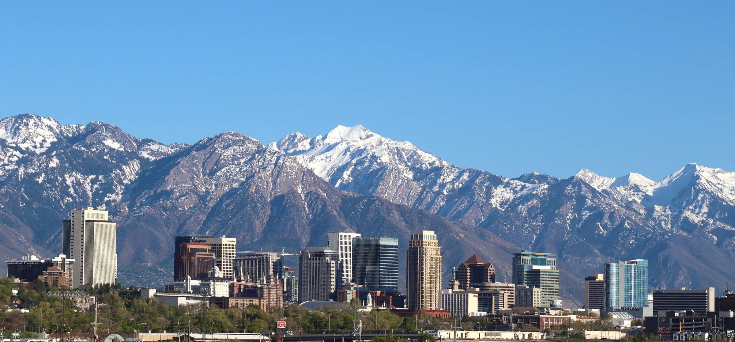Salt Lake City towers beneath snow-covered mountains, seen beyond railway tracks under a blue sky