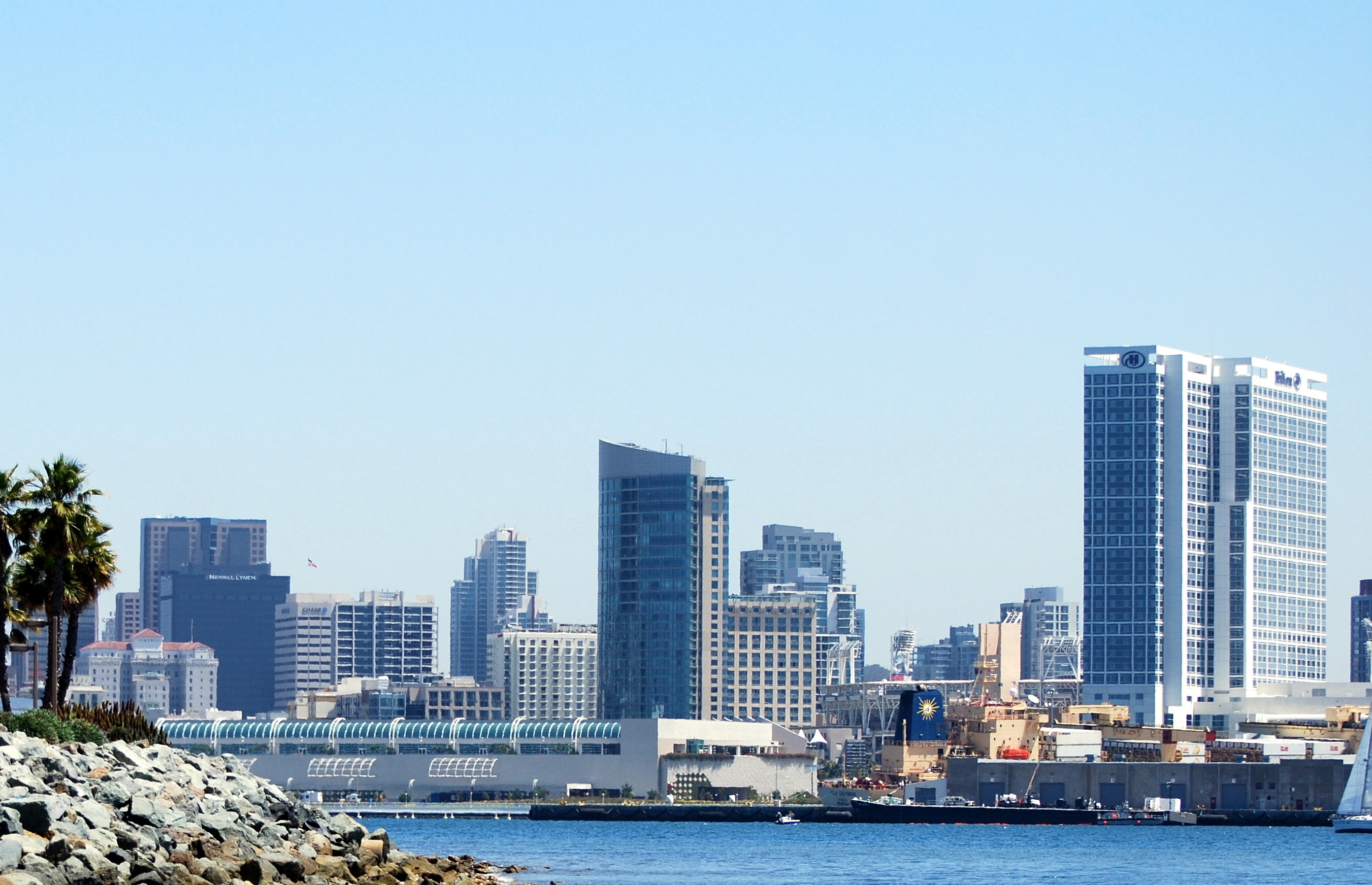 San Diego skyline across blue bay water, with rocky shore and palms at left and a ship near the waterfront