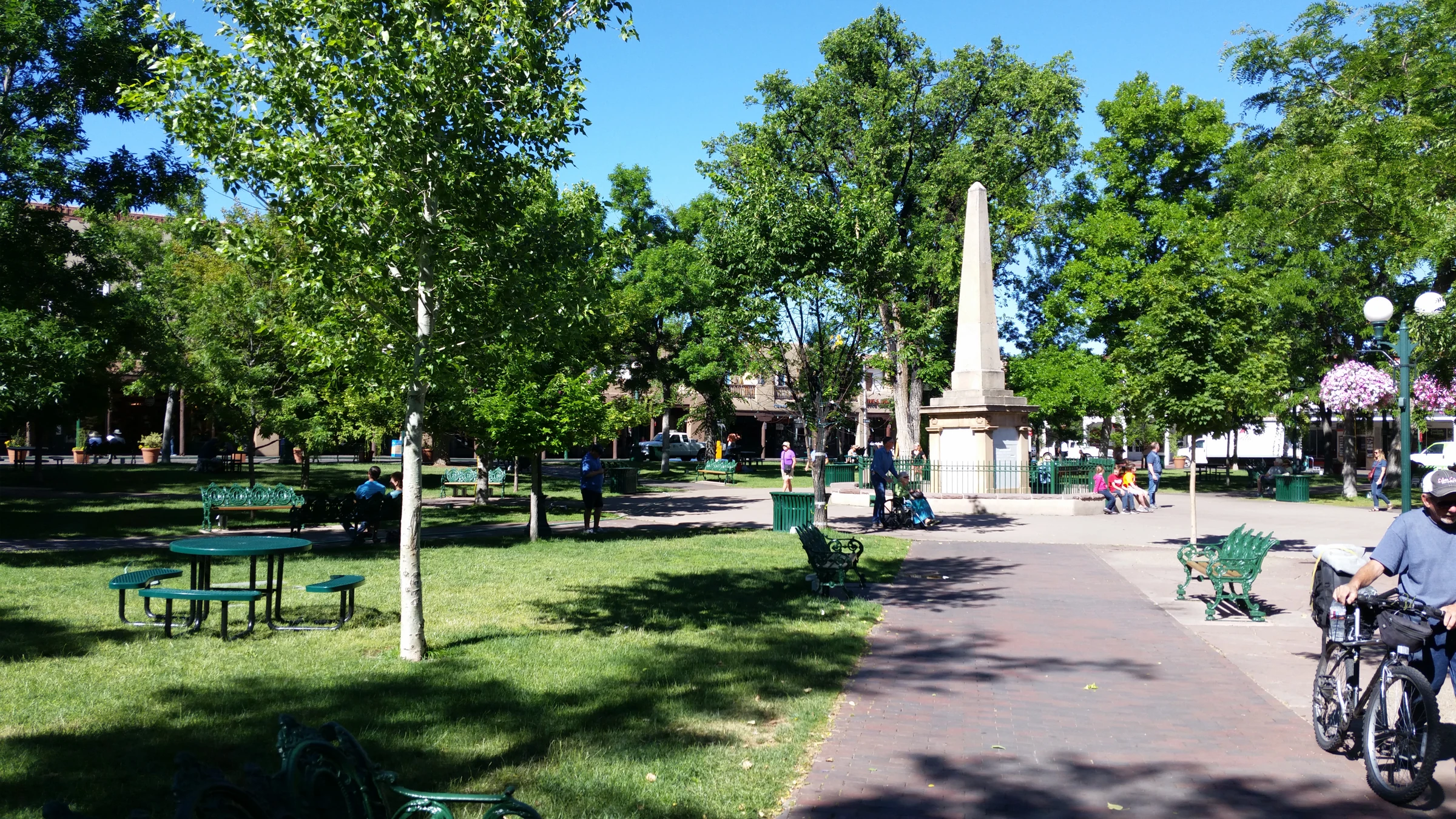 Historical Santa Fe Plaza with trees, an obelisk and shaded seating beneath blue sky