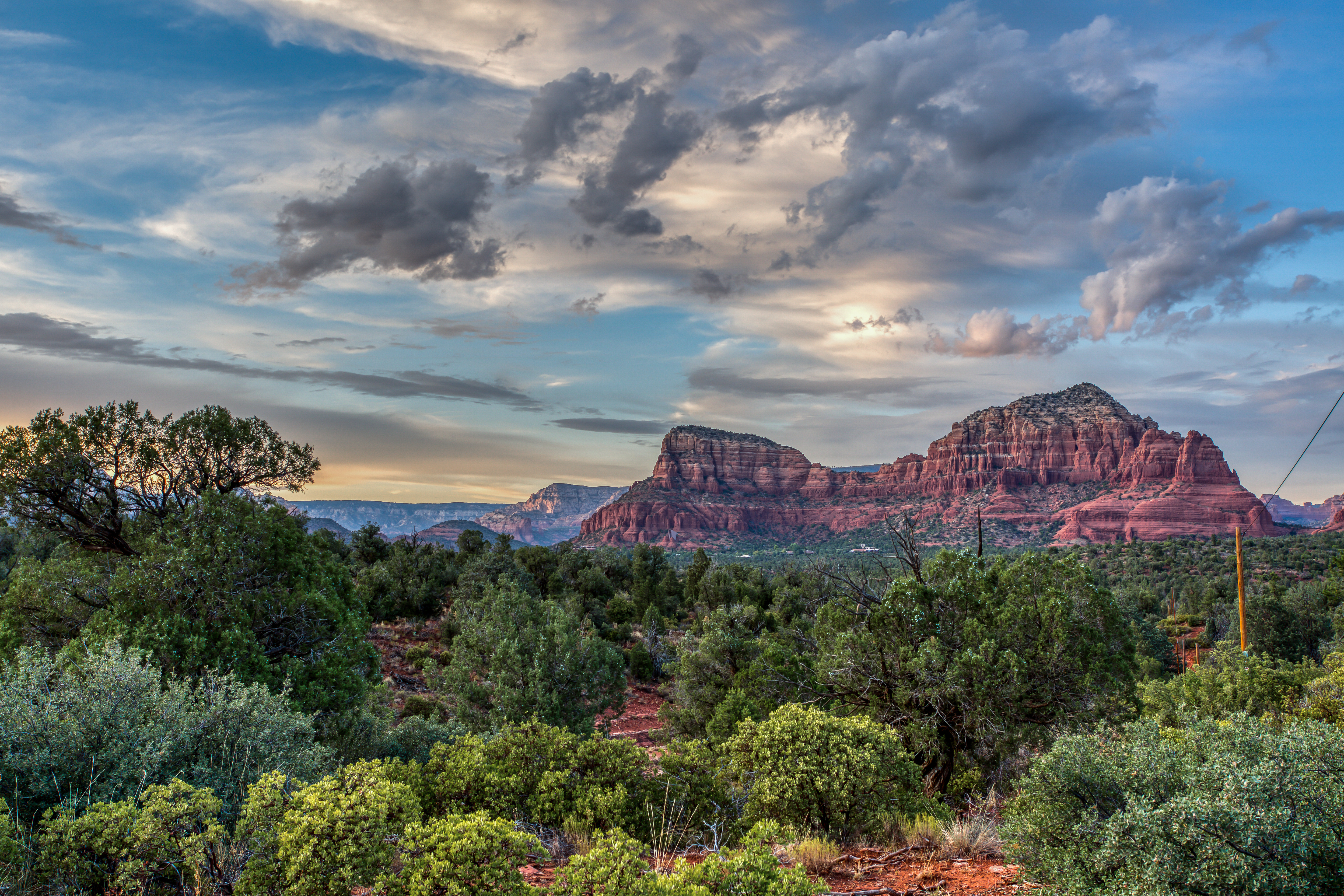 Red rock ridges above dense green scrub and trees, with a narrow earthy path beneath blue and grey cloud