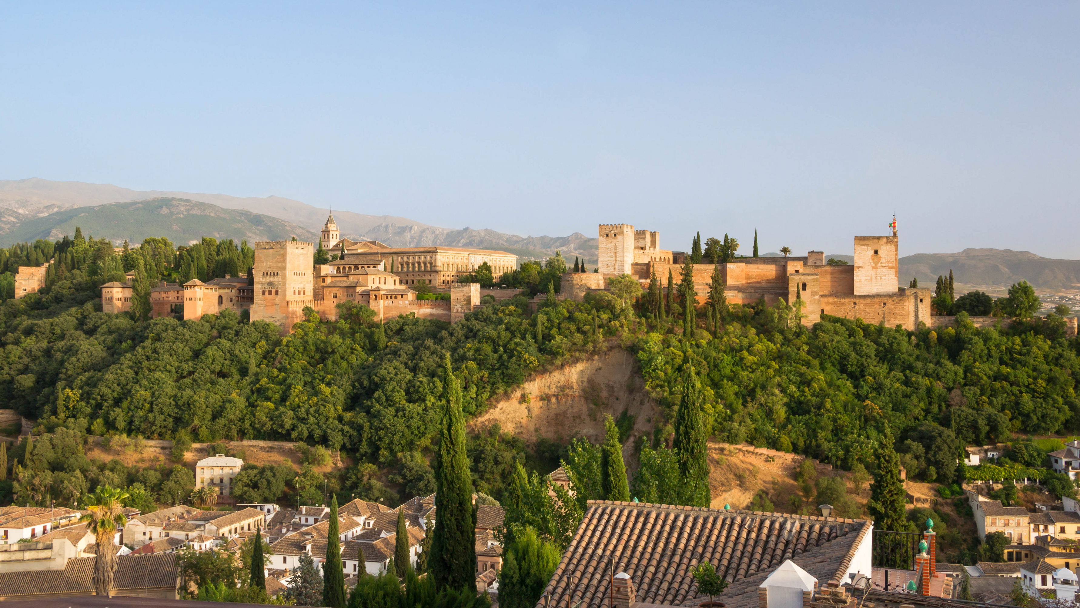 The Alhambra's stone walls and towers above a wooded slope, with hills beyond and Granada rooftops below