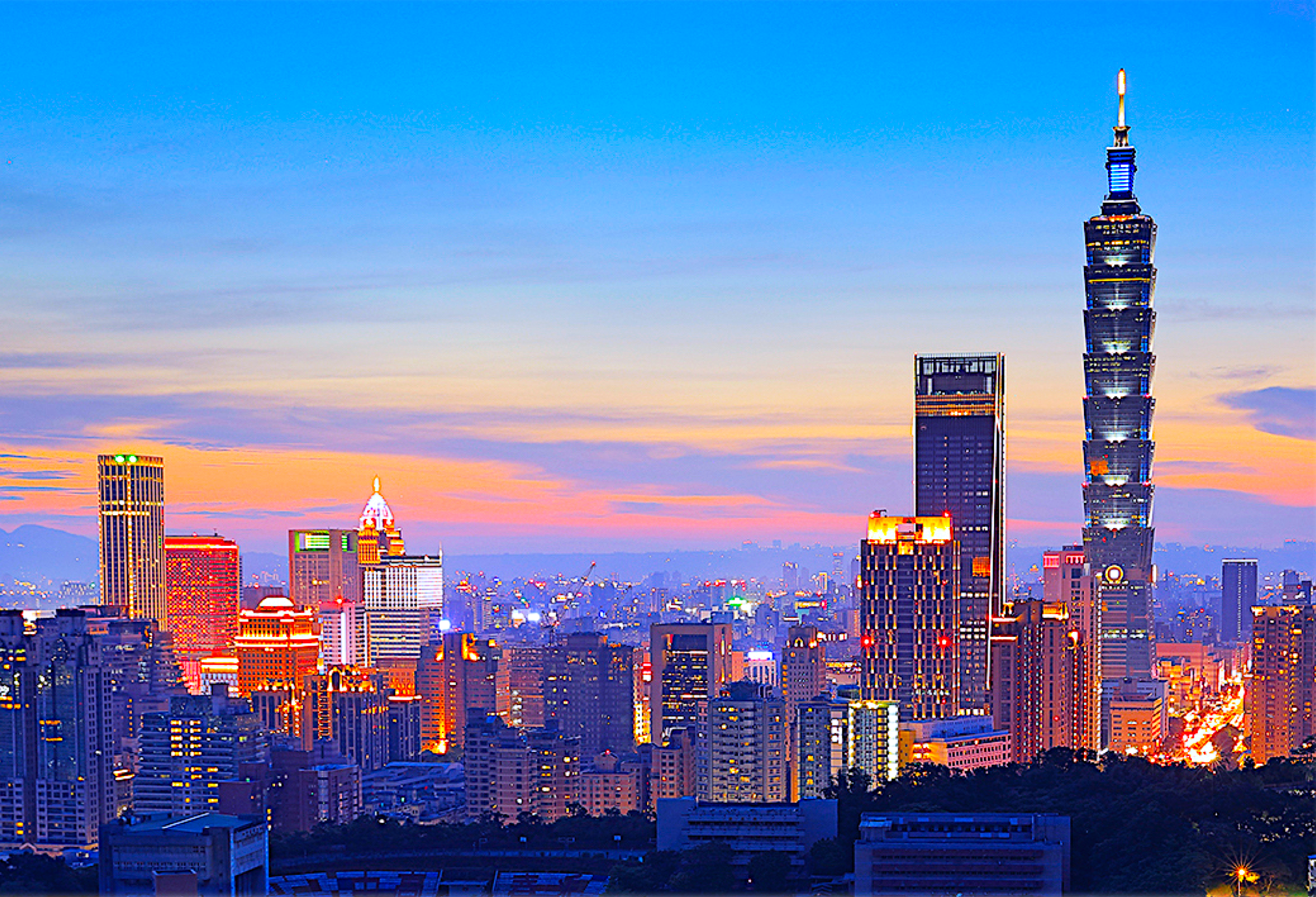 Taipei 101 at the right of a dense city skyline, with lit towers beneath a blue and pink evening sky