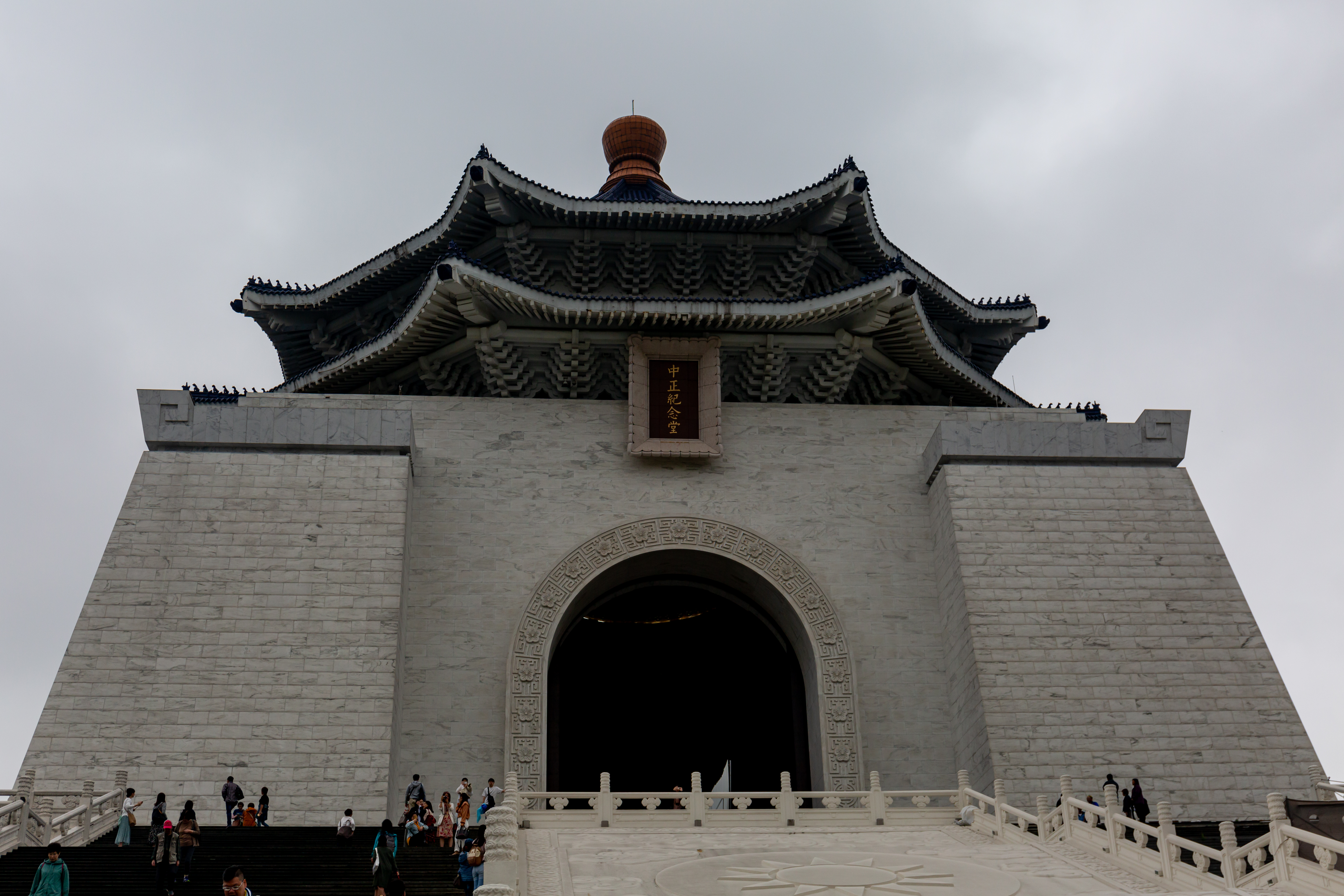 The close stone facade and blue tiled roof of Chiang Kai-shek Memorial Hall, with visitors on the steps beneath an overcast sky